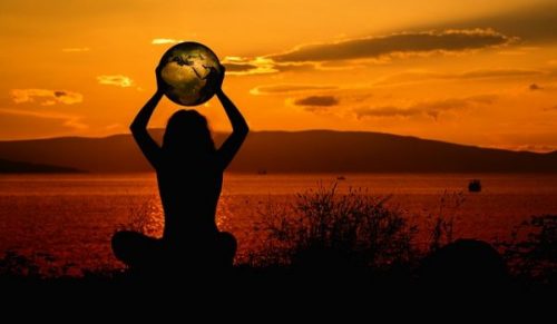 Shadowed Figure Holding Aloft A World Globe In The Sunset. Mountains, Waters And Sky In The Background.