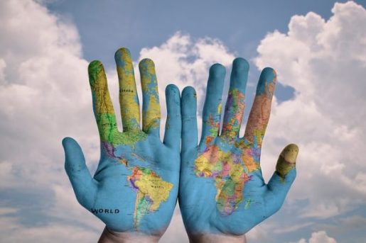 Image Of A Pair Of Open Hands Painted With A Colored World Map Backgrounded By Blue Sky, White Clouds.