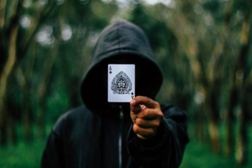 Featured Topic Image Hooded Magician Holding Up A Playing Card. The Card Is The Ace Of Spades. Out Of Focus Lined Trees In The Background.
