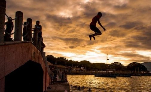 Young Adult Jumping Off Bridge/Jetty Into The Water Below. Background Sunset, Forest, Water Banks.