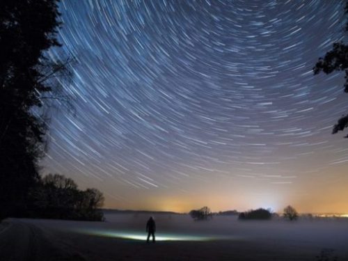 Featured Theme Image At Edge Of Forest Man In Lit Area Looks Skywards Whilst Stars Circle Overhead.