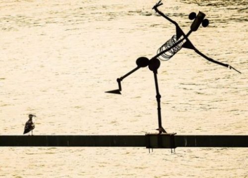 Image Of A Bird Near And Looking At A Metal Man Statue Poised Precariously Above The Waters Surface.