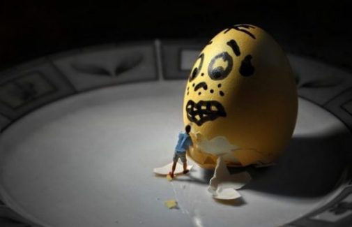 Image Of A Yellow Scared Faced Egg Watching A Tiny Toy Person Begin Deshelling.