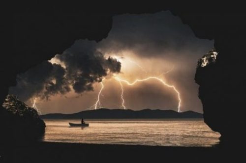 photocredit/thanks:enriquelopezgarre:pixabay Featured Image Of A Lone Man In A Boat Out At Sea While Above Storm Clouds Gather And Lightning Backgrounds The Sky.
