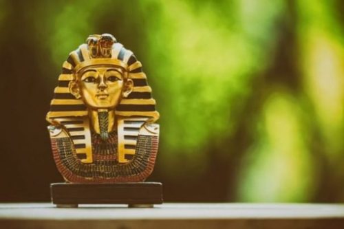 Image Of An Ancient Egyptian Mask Of A Pharaoh On A Ledge With Green Tree As Unfocused Background.